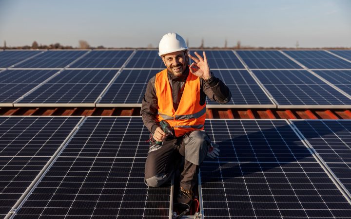 Techniker in Warnweste und Helm arbeitet auf Solarpanels und zeigt OK-Zeichen
