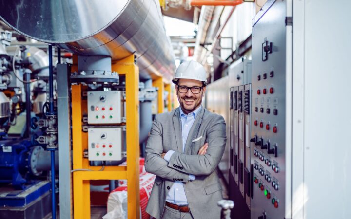 Person mit Schutzhelm und Anzug steht lächelnd in einer Industrieanlage zwischen Maschinen und Rohrleitungen.