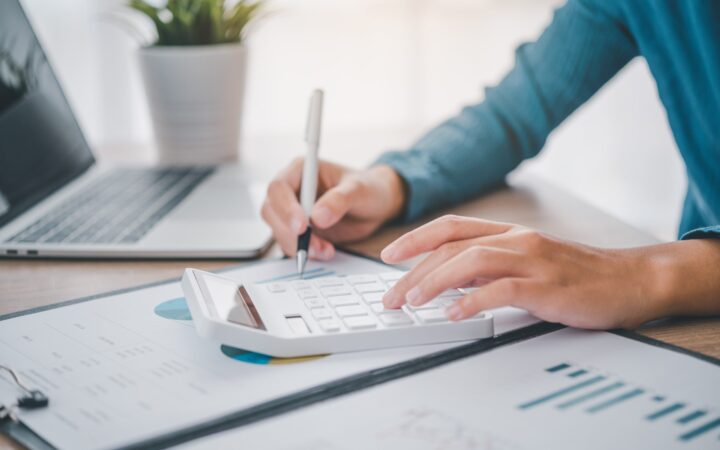 Person arbeitet am Schreibtisch mit Taschenrechner, Laptop und Dokumenten.